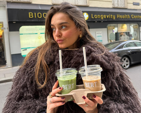 Woman holding two drinks in a city street with a building in the background